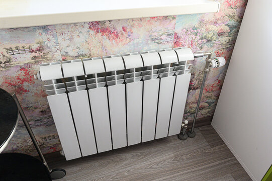 A white bimetallic radiator with 10 sections, mounted under a window; features a thermostatic valve, set against floral wallpaper, with metal pipes connected at the bottom