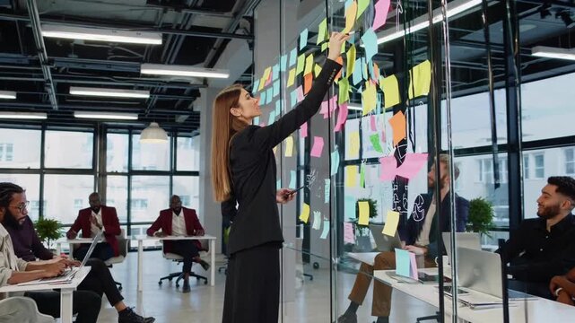 caucasian woman arranging sticky notes on glass wall during team brainstorming session with laptops and notebooks, diverse colleagues seated, colorful notes covering pane, modern open office,