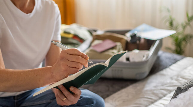 Person writing a packing checklist in a planner, preparing a suitcase for an upcoming holiday getaway, marking items and documents for a solo travel adventure and trip logistics