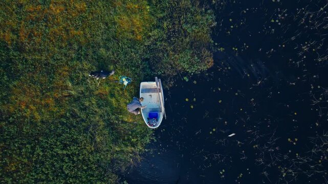 Top-down aerial shot rotating right over a man organizing gear and a blue crate in a white rowboat along a reedy shoreline with his dog.