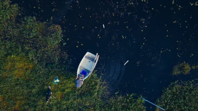 Top-down aerial shot quickly rotating left and right as a man pushes a white rowboat from a grassy shoreline into a calm body of water with his dog nearby.