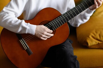 Obraz premium Close-up of man strumming guitar while sitting on yellow sofa indoors. Guitarist meditates while strumming strings. Concepts of creativity, hobbies, and music.