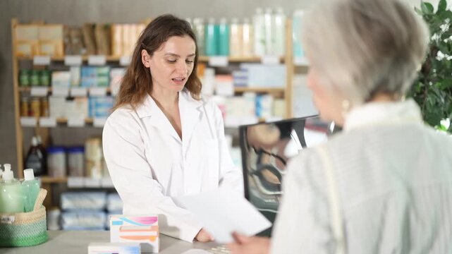 Young woman pharmacist uses a computer to check the availability of medicines and tablets in stock for a client. Elderly woman with medical prescription consults pharmacist at pharmacy. High quality