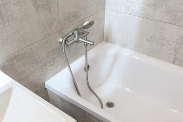 Fototapeta premium A white bathtub with a stainless steel mixer tap and flexible shower hose, set against light grey textured tiles; adjacent to a white ceramic sink.