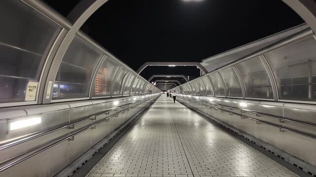 Walking perspective of a pedestrian footbridge or illuminated enclosed walkway at night in Japan. Practical walkable infrastructure enhances safety and efficency within a modern city environment