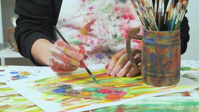 The hands of the artist, work to create creative images in art. Art paints and brushes, applying multi-coloured strokes to the canvas using palette, gouache and watercolour.