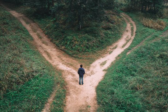 A person stands at a crossroads, two dirt paths diverging in a forest setting