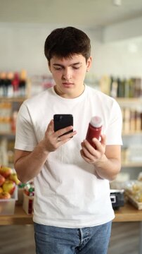 Focused interested young guy scanning barcode on colorful bottles with fresh fruit fruit smoothie using phone while choosing vitamin drinks in grocery store, checking product information and price