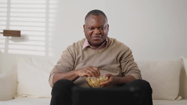 TV viewer relaxing in front of television set and eating popcorn in weekend. Mature african american man spending evening at home, enjoying favorite series or sport match, entertainment and leisure
