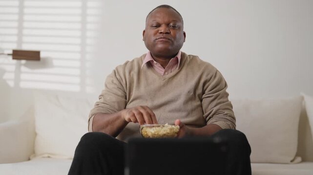 Lazy weekend at home, mature black man watching TV and eating popcorn, portrait. Entertainment and leisure in evening at home, middle-aged african american male person enjoying TV show or sport match
