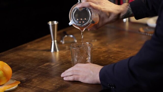 Bartender serving an old fashioned cocktail with a strainer into a glass with a big ice rock.