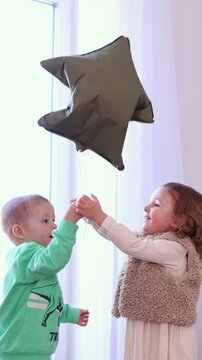 A young boy in a turquoise hoodie and a little girl in a fluffy beige vest are reaching their hands up towards a large, olive-green star-shaped balloon. 