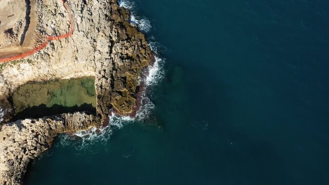 Aerial view of rugged rocky coastline meeting the deep blue sea in Polignano a Mare, Italy. Clear water and textured cliffs create a dramatic natural landscape.