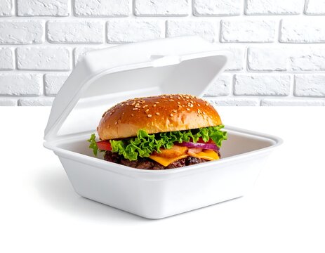 A sesame seed topped burger rests in a white styrofoam takeout container against a white brick background