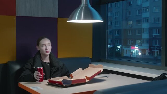 A young woman sits alone in a modern pizzeria at night, eating pizza and drinking a soda. She looks thoughtfully out the window before turning and taking a sip. Urban city view in background.