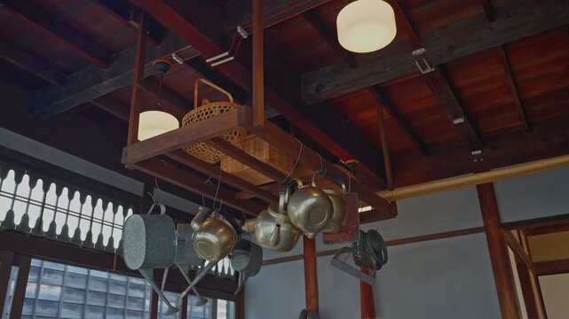 A cinematic low-angle shot of overhead storage in a historic Japanese merchant house. Features brass kettles and bamboo baskets hanging from dark wooden beams at Edo-Tokyo Open Air Architectural Museu