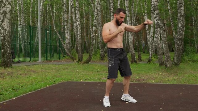 A fit, shirtless man with a beard shadowboxes on a sports court in a birch forest. He practices punches and footwork. The camera moves closer for an intense, dynamic shot of him punching.