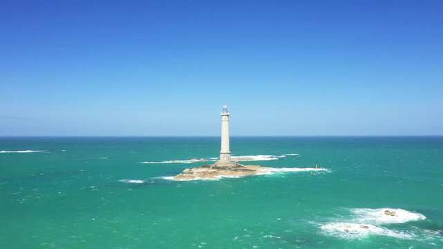 A solitary lighthouse stands on a rocky islet surrounded by turquoise sea under a clear blue sky at Cap de la Hague, Normandy, France.