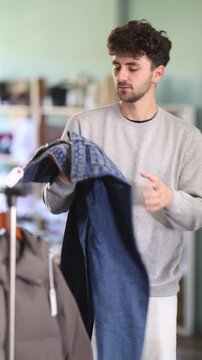 Regular customer guy client pre-examine fabric and style of product, is busy choosing jeans trousers. Client enjoys shopping for clothes at store browsing through colorful on display racks