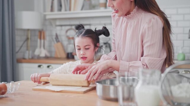 Asian mother guides her young daughter as they use a wooden rolling pin to flatten dough together, enjoying a creative baking day in their bright modern home kitchen.