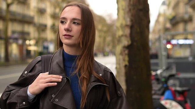 A woman in a leather jacket stands on a city street in Paris, adjusting a bag strap on her shoulder while holding a paper coffee cup.