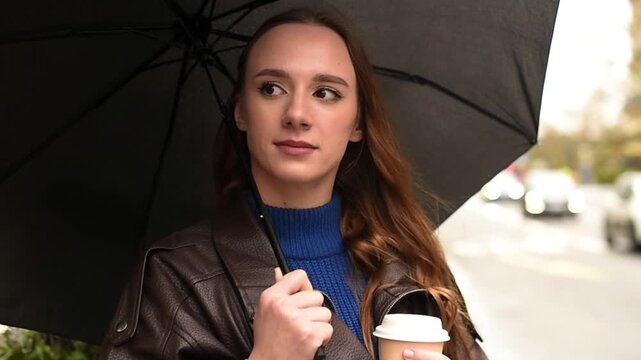 A woman in a leather jacket stands on a street in Paris under a black umbrella while holding a paper coffee cup on a rainy day.