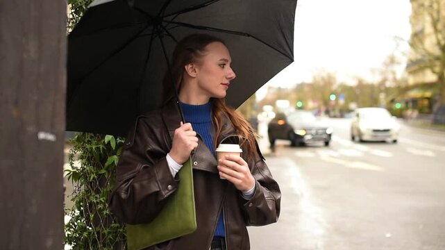A woman in a leather jacket stands on a Paris street under a black umbrella, holding a paper coffee cup on a rainy day.