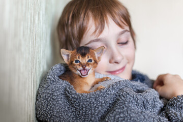 Close-up of orange kitten meowing while nestled inside hood of boy’s cozy fleece jacket. Warm indoor portrait, soft textures, companionship and tender moment between child and pet. © Natallia