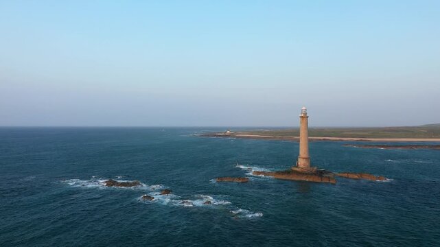 A tall lighthouse stands on rocky outcrops surrounded by the sea at Cap de la Hague, Normandy, France under a clear sky. Rugged coastline and open water create a dramatic maritime landscape.