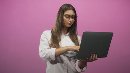 Woman holding laptop and typing with hands in a pink studio, looking at screen with pursed lips; concentration remote work productivity.