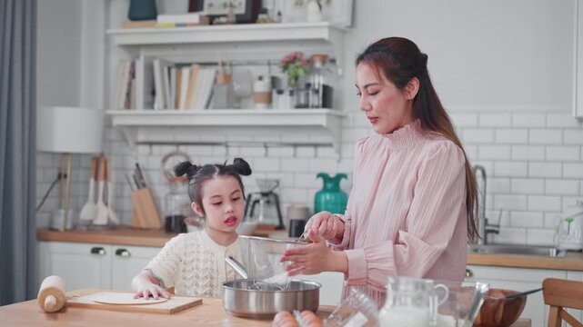 Loving Asian mother helps her little daughter sift flour while baking together in a bright modern kitchen, enjoying a fun and educational family morning.