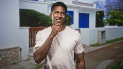Fototapeta premium Black man touching chin and smiling on a street in front of white building with blue door; thoughtful curiosity.
