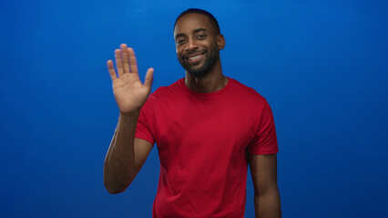 Man smiling and waving hand in blue studio, palm facing camera, wearing red shirt and gold chain  welcoming greeting. © Krakenimages.com