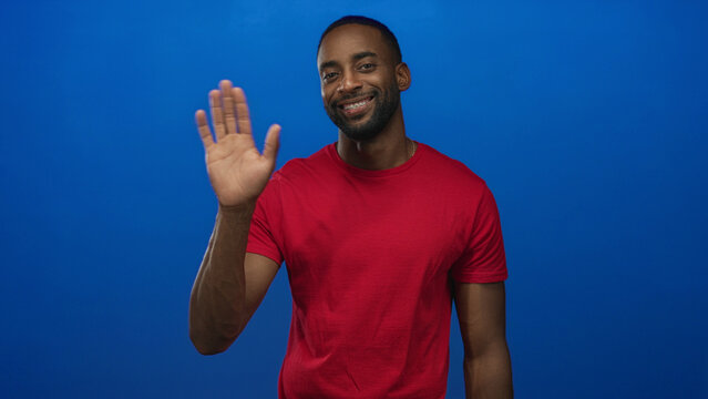 Man smiling and waving hand in blue studio, palm facing camera, wearing red shirt and gold chain; welcoming greeting.