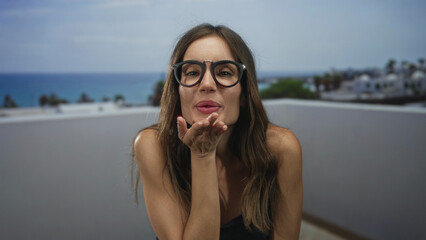 Fototapeta premium Woman blows a kiss through black glasses with both hands on a seaside balcony overlooking the ocean; affection longing.