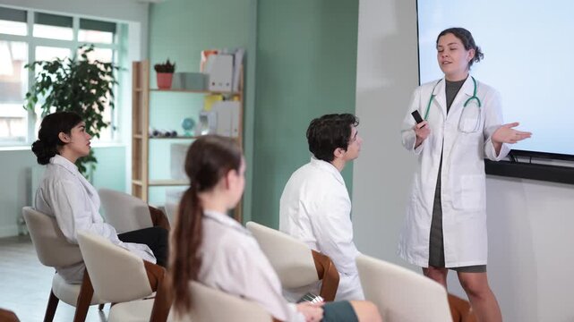 Positive young girl intern standing near interactive board telling new topic or showing presentation with remote control to medical students sitting on chairs in light classroom