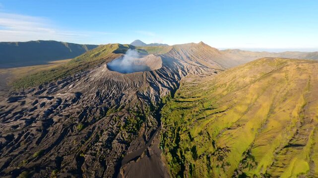 Cinematic aerial view of active Mount Bromo volcano emitting smoke with Mount Semeru in the distance at sunrise. Dramatic volcanic landscape in East Java, Indonesia.