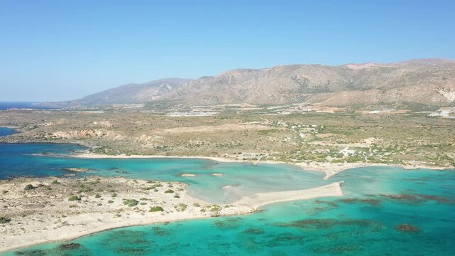 Stunning aerial view of Elafonisi beach with clear turquoise waters, sandy shores, and rugged mountains in Crete, Greece. The landscape features a unique sandbar and natural coastal beauty.