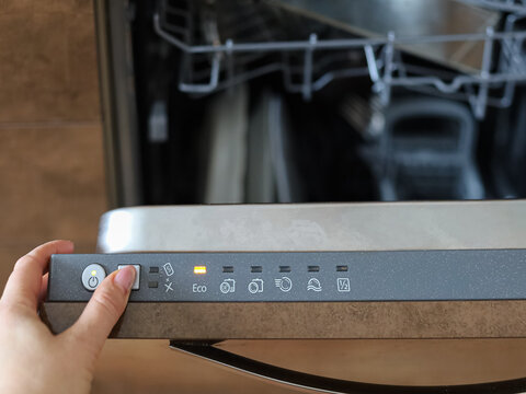 Hand operating dishwasher control buttons with indicator lights on, concept of smart kitchen technology and automated cleaning.