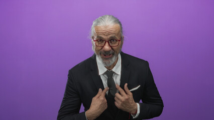 Man in suit adjusting tie and pointing to chest in studio, wearing glasses and grey beard while...