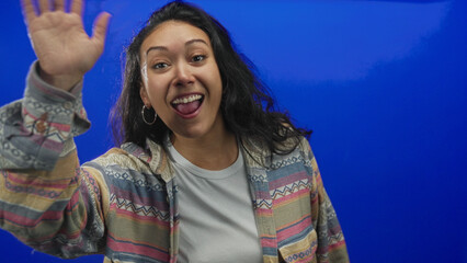 Woman waving hand toward camera in blue studio wearing patterned shirt and hoop earring  joyful greeting. © Krakenimages.com