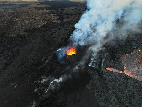 Aerial view of fiery lava flows contrast sharply against the stark black volcanic landscape, with plumes of smoke rising into the sky, Grindavik, Iceland.