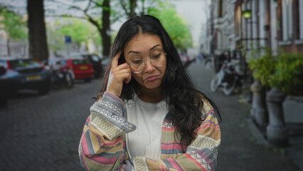 Fototapeta premium Woman finger to temple on a cobblestone city street wearing round glasses and patterned jacket, arms crossed loosely; doubt thinking contemplation.