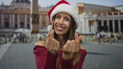 Fototapeta premium Young hispanic woman in a santa hat makes a finger heart gesture outside a historic building; romance.