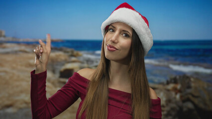 Fototapeta premium Woman wearing santa hat and burgundy top pointing finger to sky at sunny beach shore under clear blue sky; anticipation.