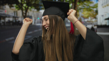 Fototapeta premium Young woman in academic gown dancing with raised fists on bustling city street; achievement moment celebration joy.