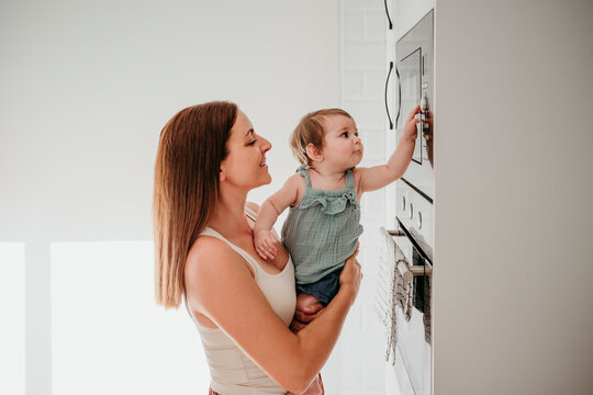 Mother holding baby girl pressing microwave button in home kitchen