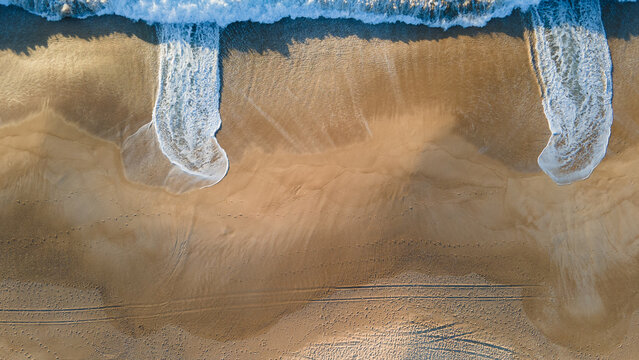 Aerial view of the golden sand meeting the turbulent, frothy waves of the Atlantic, creating a mesmerizing dance of textures and tones, Nazare, Leiria District, Portugal.