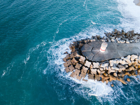 Aerial view of a lighthouse standing firm against the turquoise ocean waves crashing on the shore, Nazare, Leiria District, Portugal.