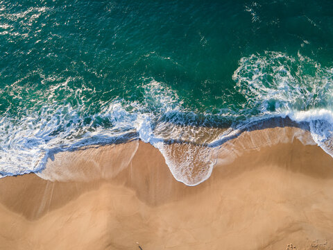Aerial view of turquoise waves crashing onto the golden sands of the beach, a stunning contrast of color and texture, Nazare, Leiria District, Portugal.